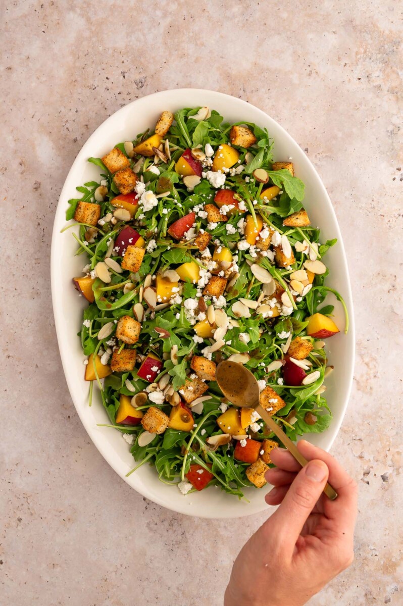 A hand drizzling balsamic dressing over peach salad with goat cheese on a large white platter.