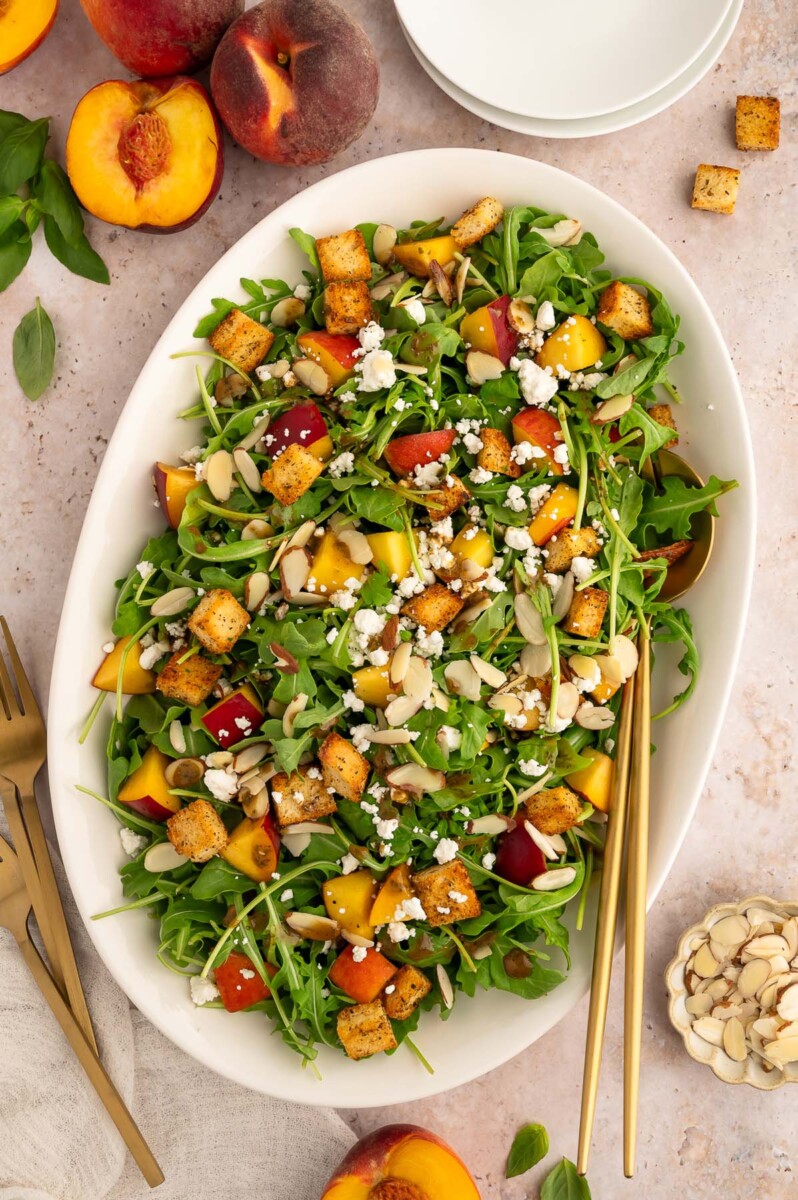 Peach Salad with goat cheese on a large white platter with gold utensils in it and fresh peaches on the side.
