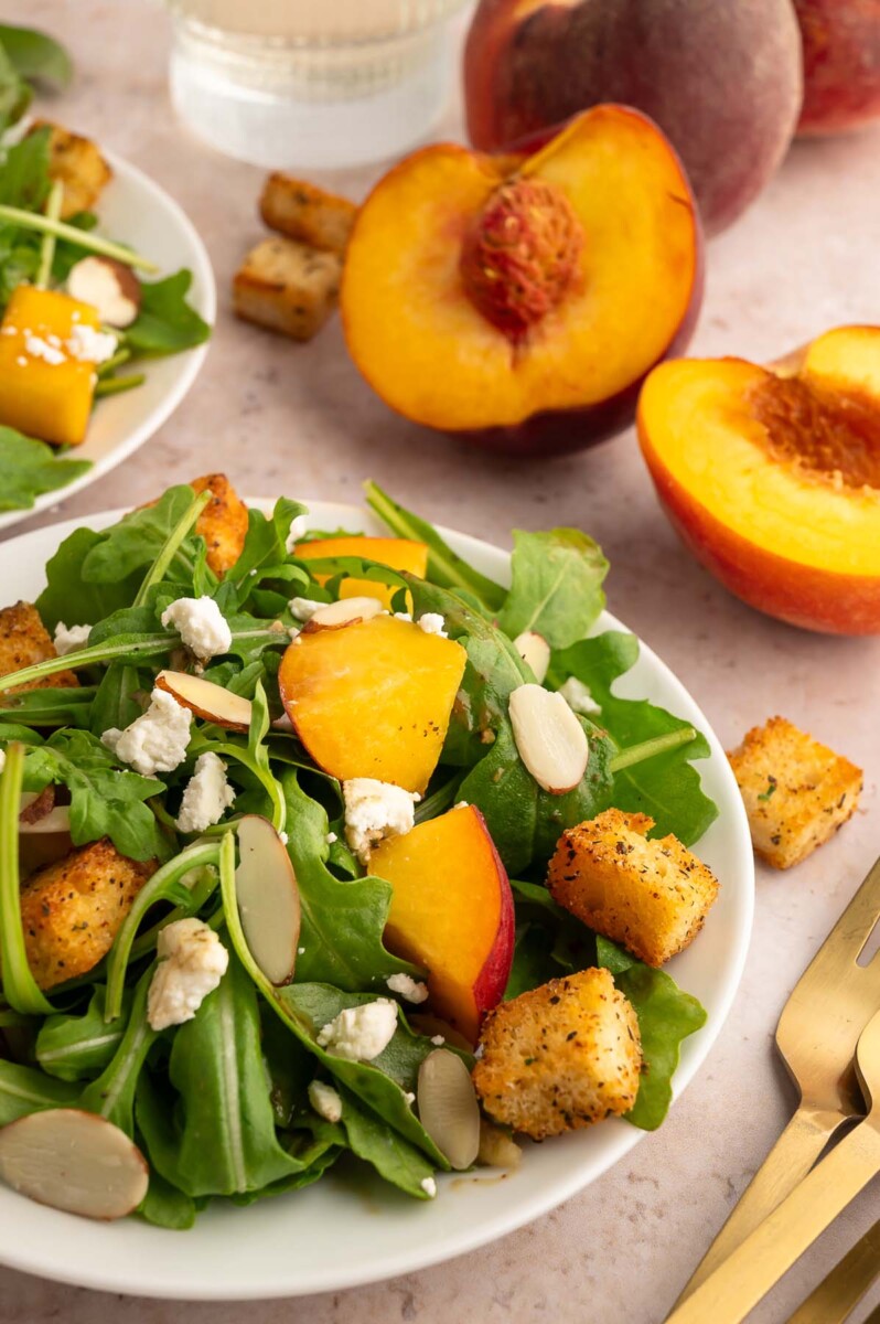A simple peach salad topped with homemade croutons and sliced almonds with fresh peaches in the background.