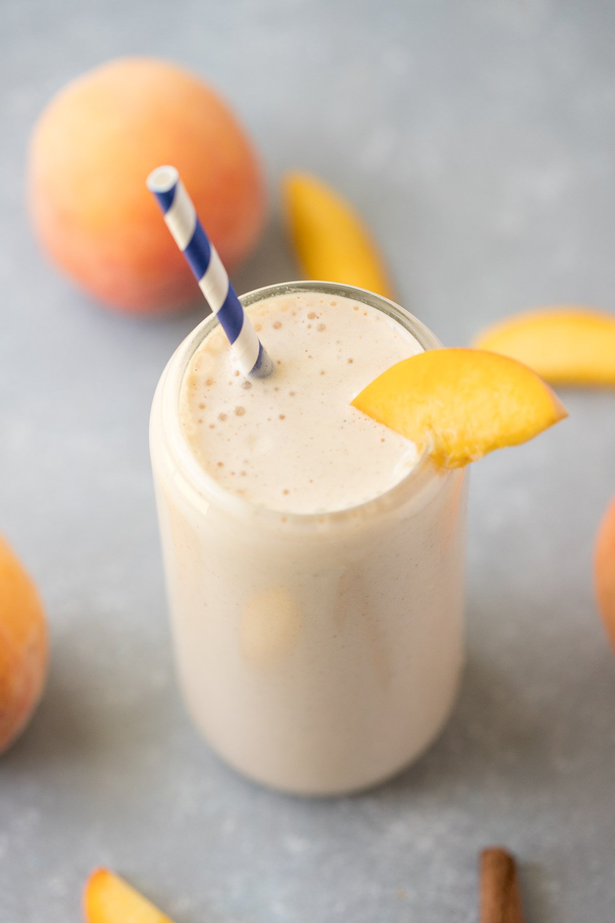 A peach smoothie in a clear glass with a blue and white striped straw in it garnished with a peach slice and more sliced peaches in the background