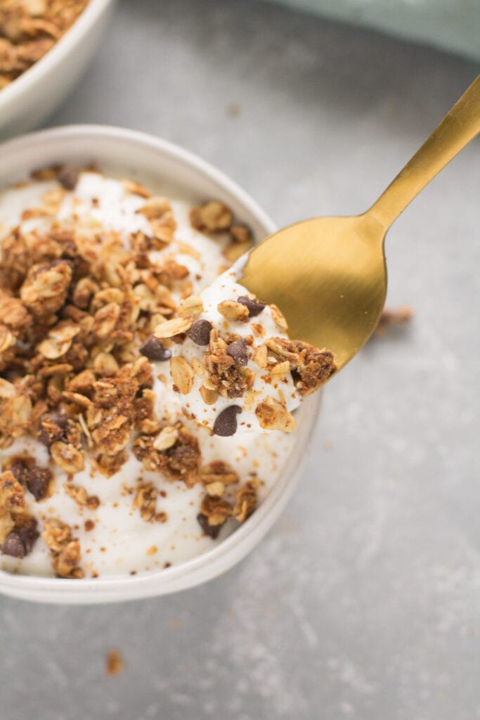 Peanut Butter Granola on a bowl of plain greek yogurt with a gold spoon taking a bite out of it.