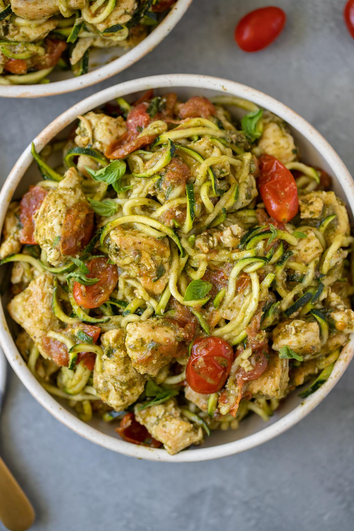 Whole30 Pesto Chicken Zucchini Noodles in a white bowl with tomatoes and basil scattered around.
