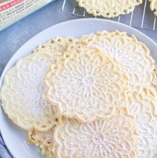 Pizzelle Cookies on a white plate with a cooling rack and back of flour off to the side.
