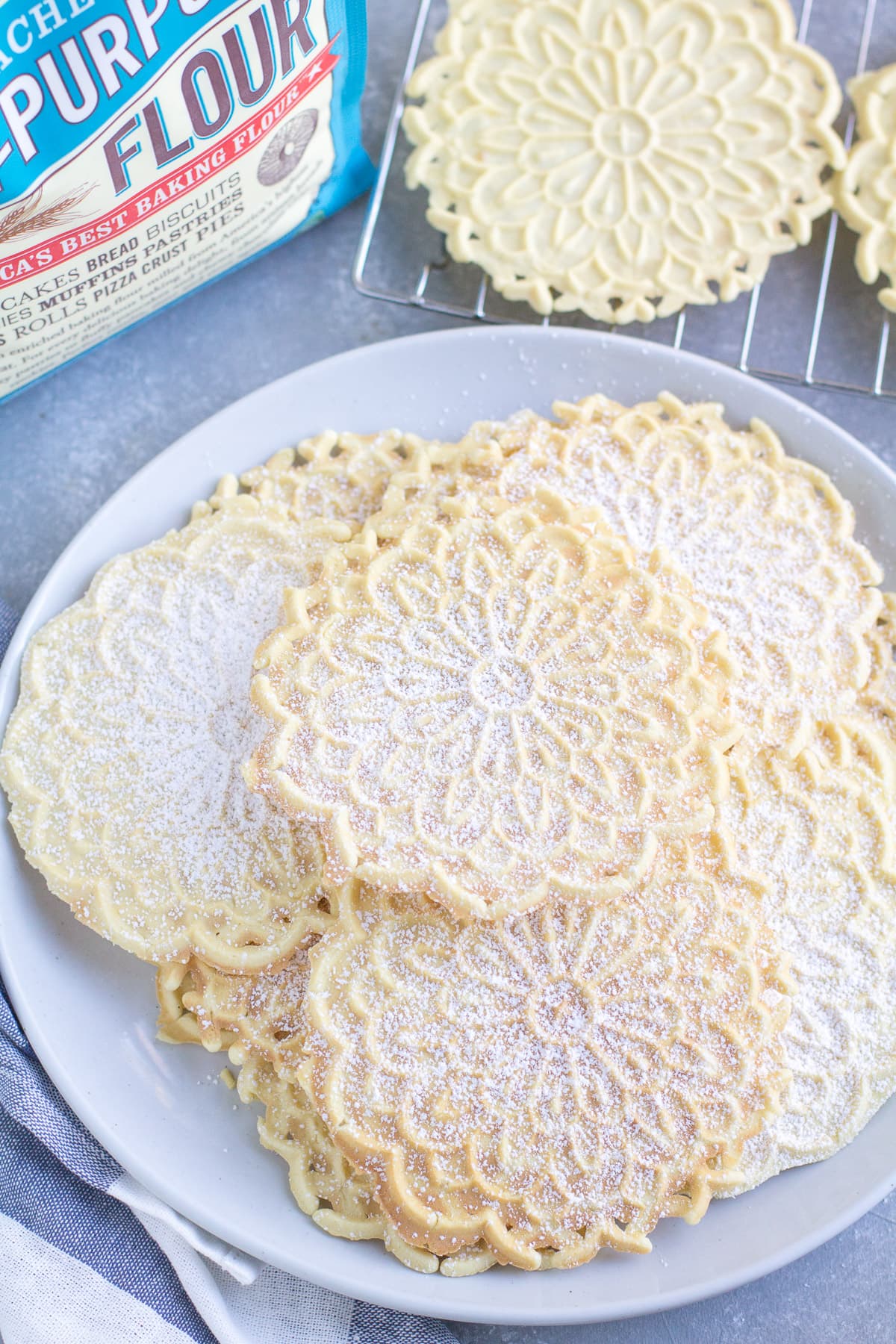 Pizzelle Cookies on a white plate with a cooling rack and back of flour off to the side.