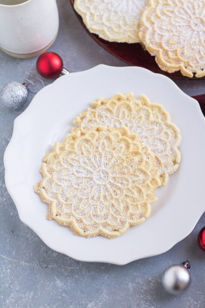 Pizzelle on a plate with cookies off to the side and a cup of coffee/holiday ornaments in the background.