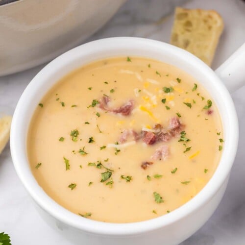 A white bowl of potato leek soup topped with bacon bits and cheese on a grey background with a dutch oven on the side