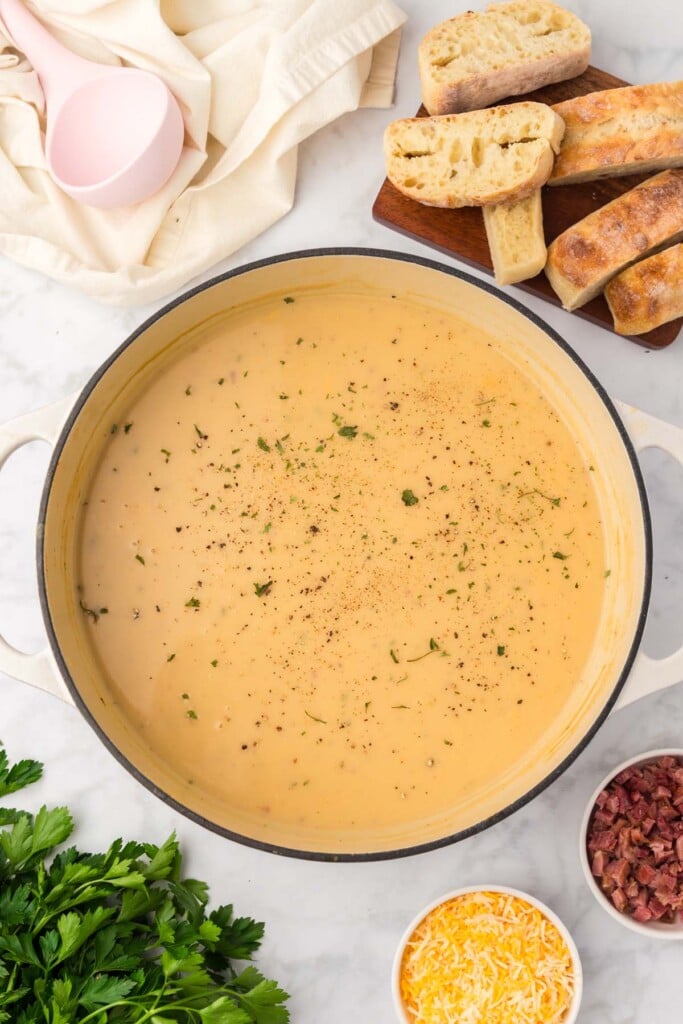 An overhead shot of potato leek soup in a dutch oven with a ladle, crusty bread, cheese and bacon bits on the side. 