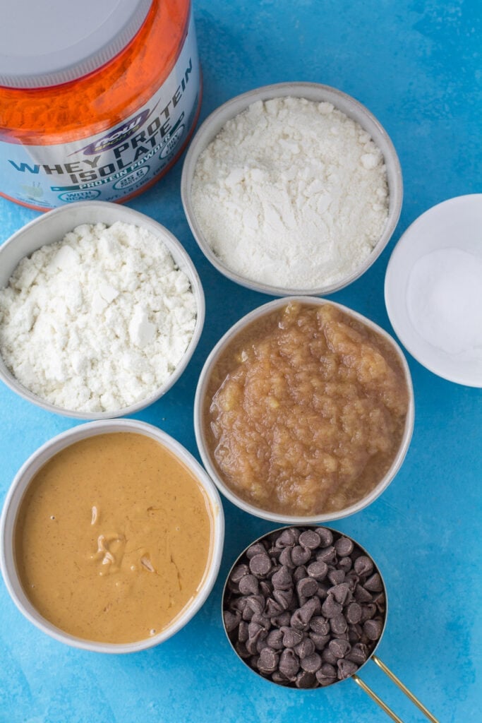 Ingredients for protein cookies in small white bowls: peanut butter, chocolate chips, applesauce, protein powder, coconut flour and baking soda.