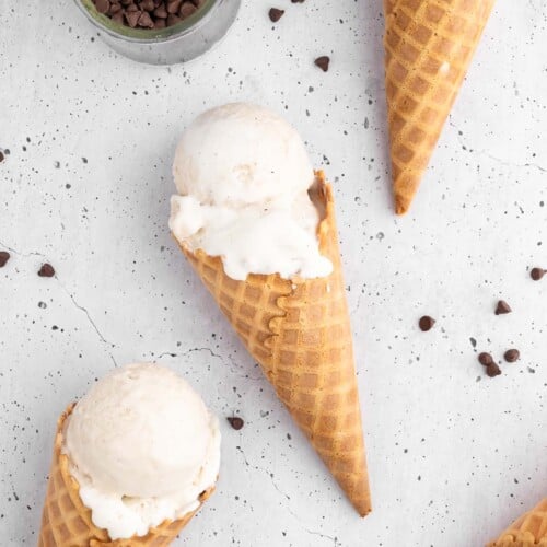 Vanilla protein ice cream in waffle cones on a white background with mini chocolate chips scattered around and a small bowl of them on the side