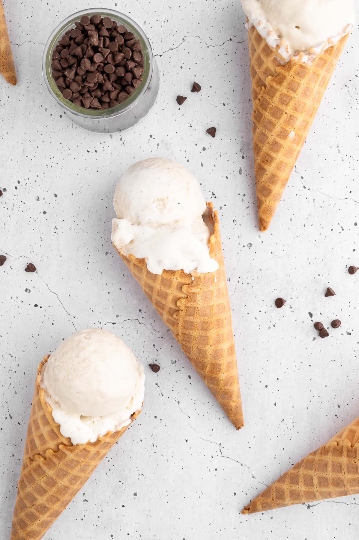 Vanilla protein ice cream in waffle cones on a white background with mini chocolate chips scattered around and a small bowl of them on the side