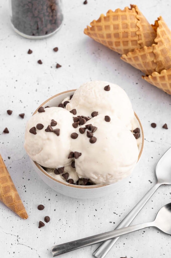 Three scoops of vanilla protein ice cream in a bowl topped with mini chocolate chips with more chocolate chips, waffle cones, and spoons on the side
