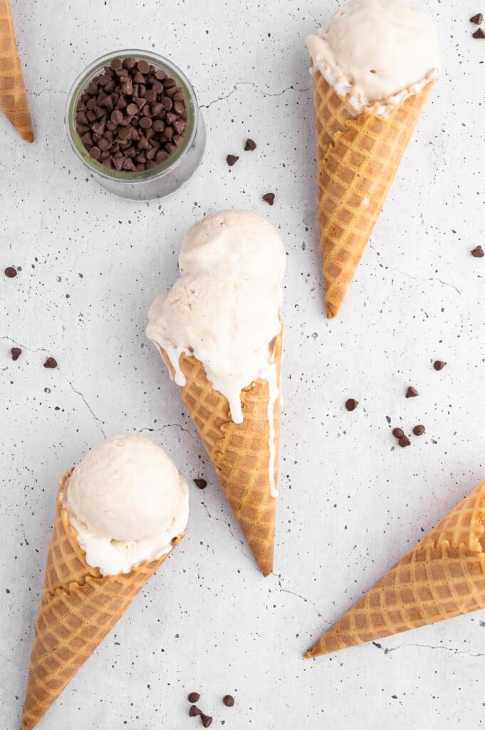 Vanilla protein ice cream scoops in waffle cones on a white background with mini chocolate chips scattered around and a small bowl of them on the side