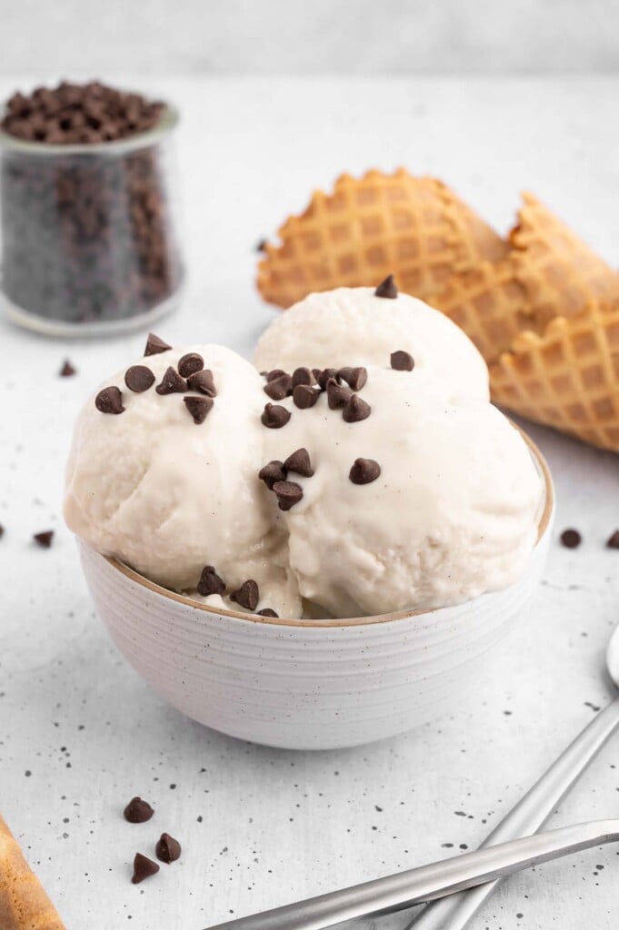 Three scoops of vanilla protein ice cream in a bowl topped with mini chocolate chips with more chocolate chips, waffle cones, and spoons on the side