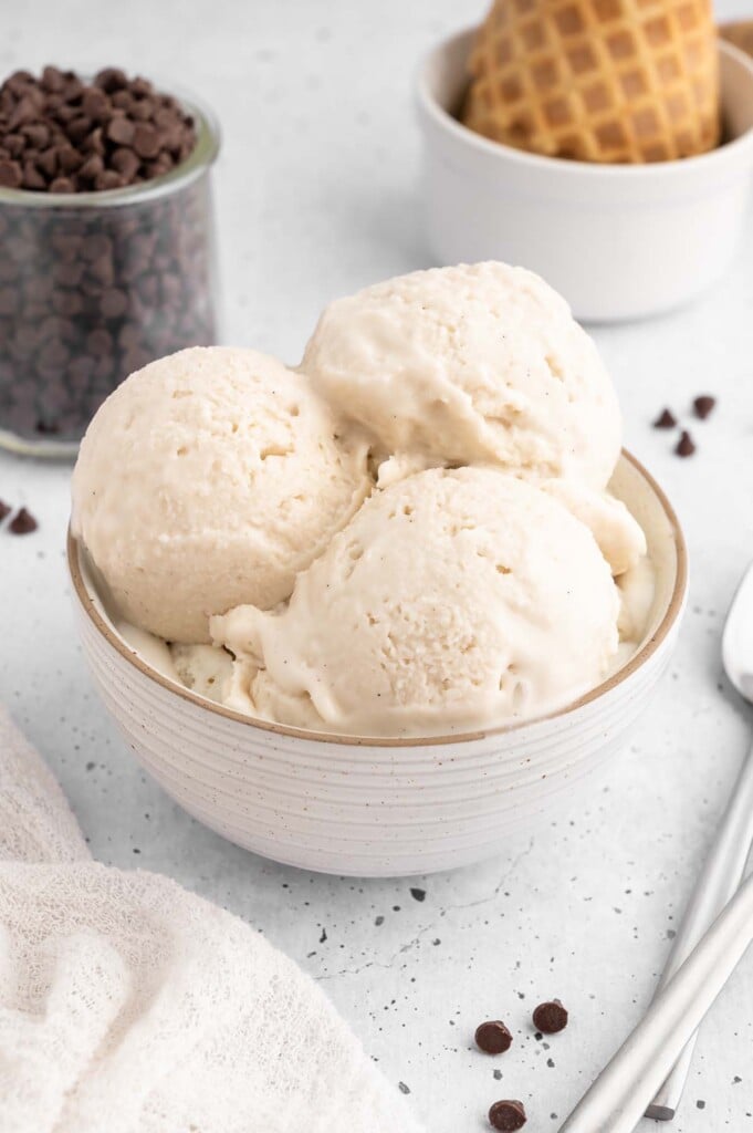 Three vanilla protein ice cream scoops in a white bowl with chocolate chips in a small bowl and scattered on the white counter with waffle cones in the background