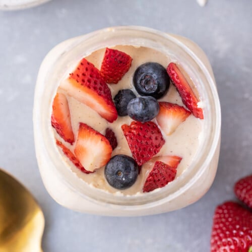 An overhead shot of protein overnight oats with strawberries and blueberries on top with a gold spoon on the side with a grey background