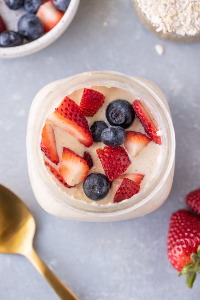 An overhead shot of protein overnight oats with strawberries and blueberries on top with a gold spoon on the side with a grey background