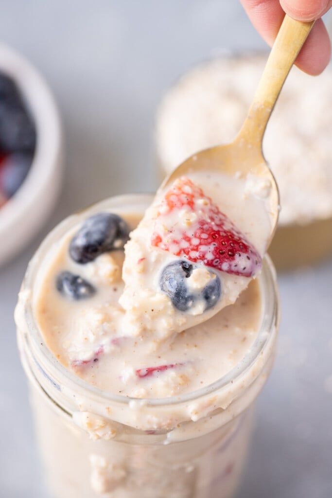 A gold spoon taking a large bite out of a mason jar of protein overnight oats topped with strawberries and blueberries