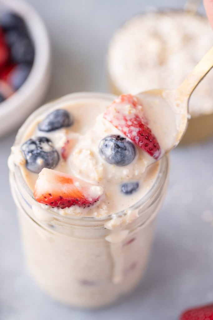 A gold spoon taking a large bite out of a mason jar of protein overnight oats topped with strawberries and blueberries