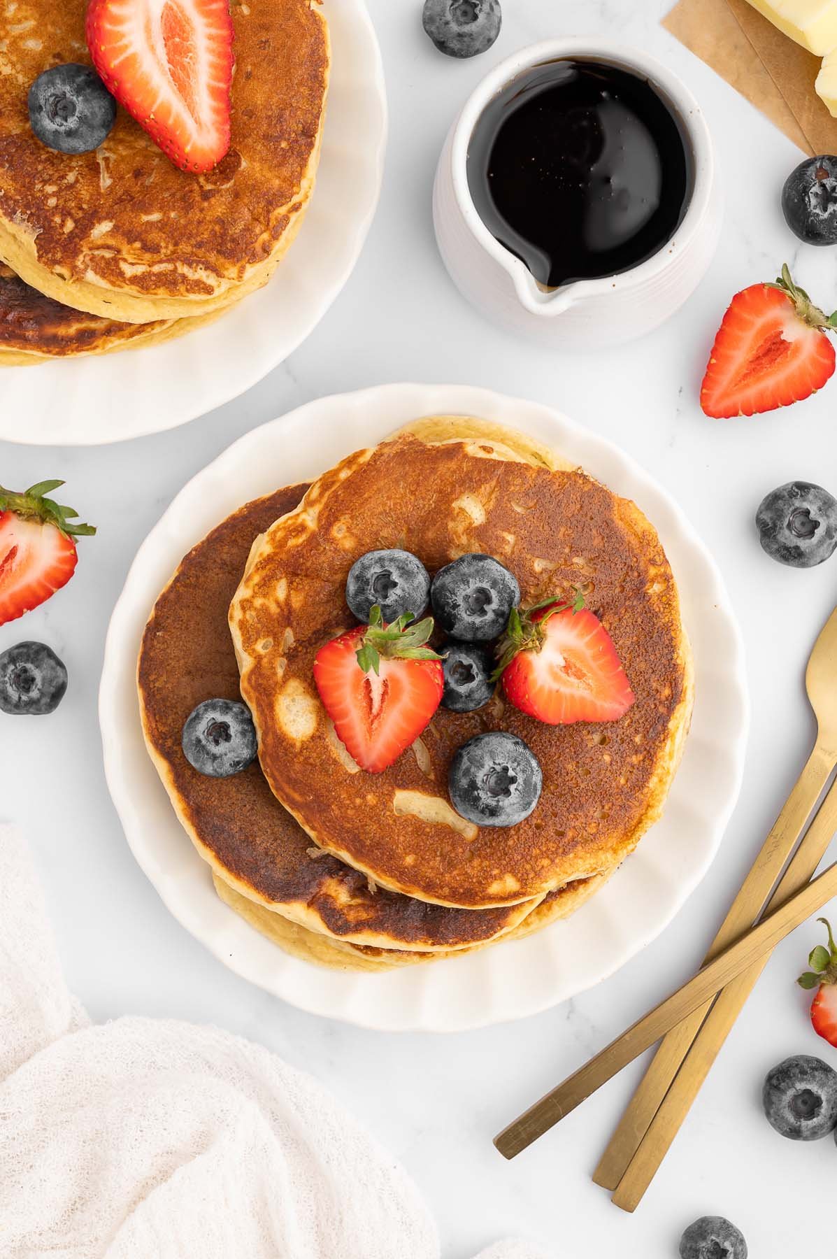 Protein Pancakes stacked on a white plate with strawberries and blueberries on top with another plate of pancakes, fruit and a small white pitcher of maple syrup on the side.