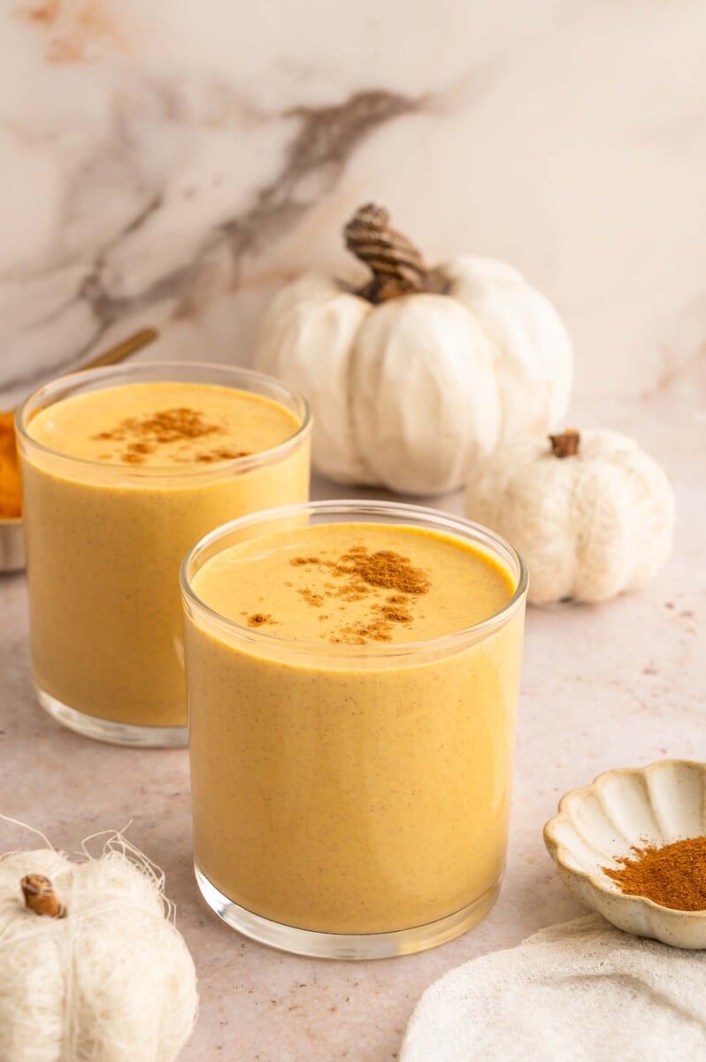 Two clear glasses of pumpkin protein smoothie topped with pumpkin pie spice with white pumpkins in the background.