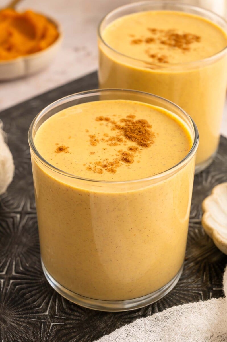 Pumpkin protein smoothie topped with pumpkin pie spice in a clear glass with another smoothie in the background.