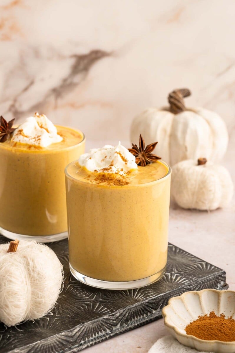 2 pumpkin smoothies in clear glasses topped with pumpkin pie spice, whipped cream, and Star anise garnish with white pumpkins in the background.