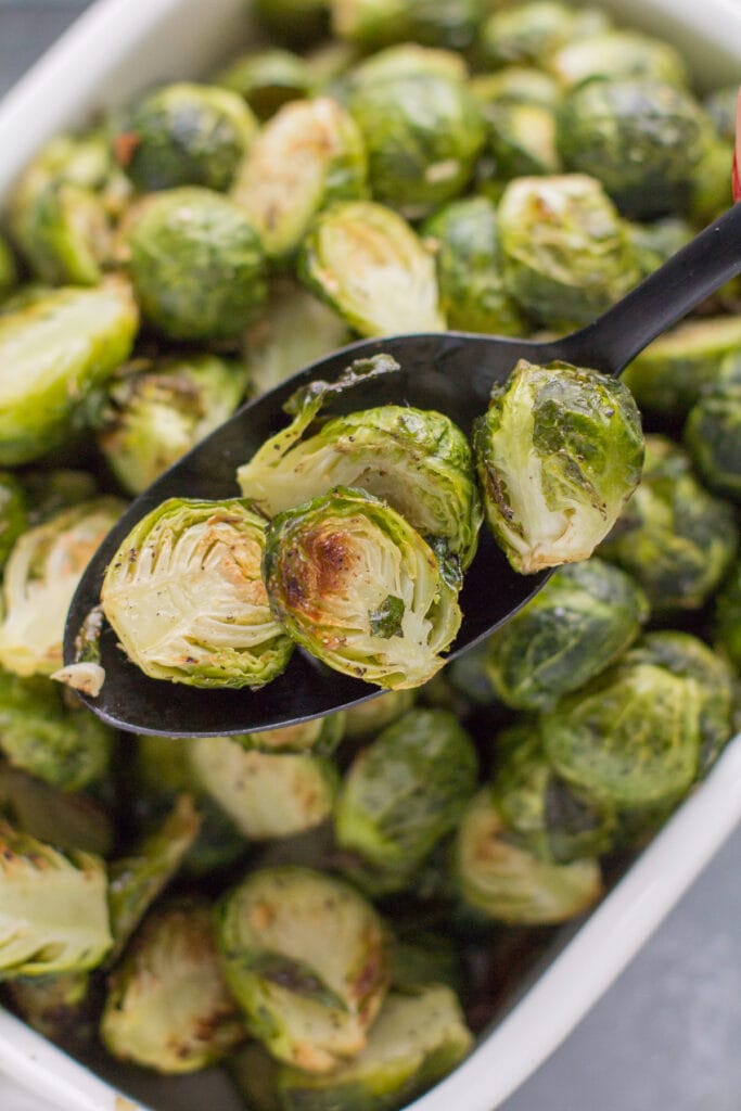 Roasted Brussels Sprouts on a black serving spoon taking a scoop out of a white serving bowl.