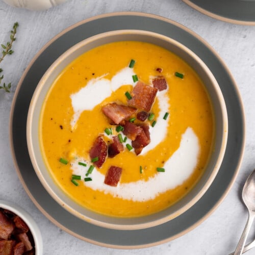 An overhead shot of roasted butternut squash soup in a grey bowl on a grey background with bacon and chives on top
