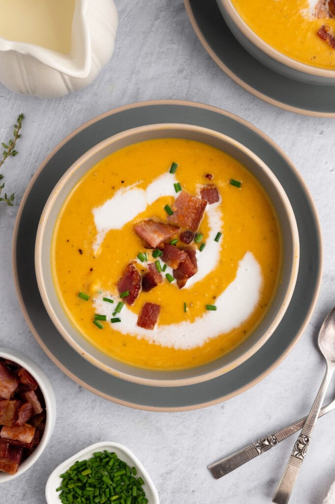 An overhead shot of butternut squash soup recipe in a grey bowl on a grey background with bacon and chives on top
