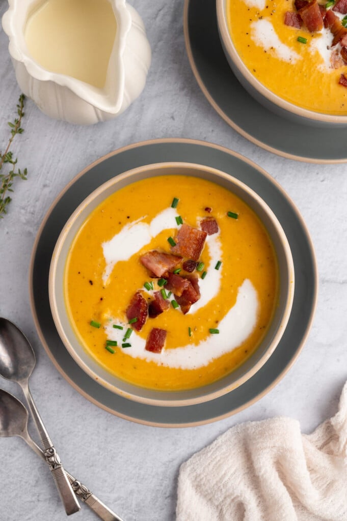 An overhead shot of roasted butternut squash soup in a grey bowl on a grey background with another bowl on the side with bacon and chives on top