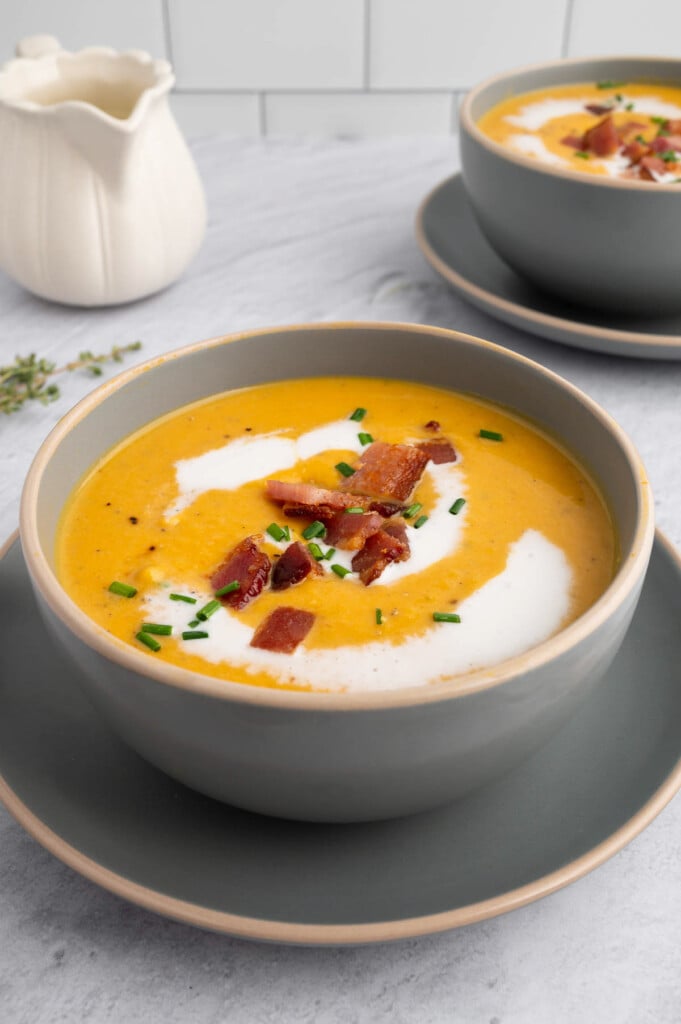A grey bowl of roasted butternut squash soup with chives and bacon on top and another bowl of soup in the background