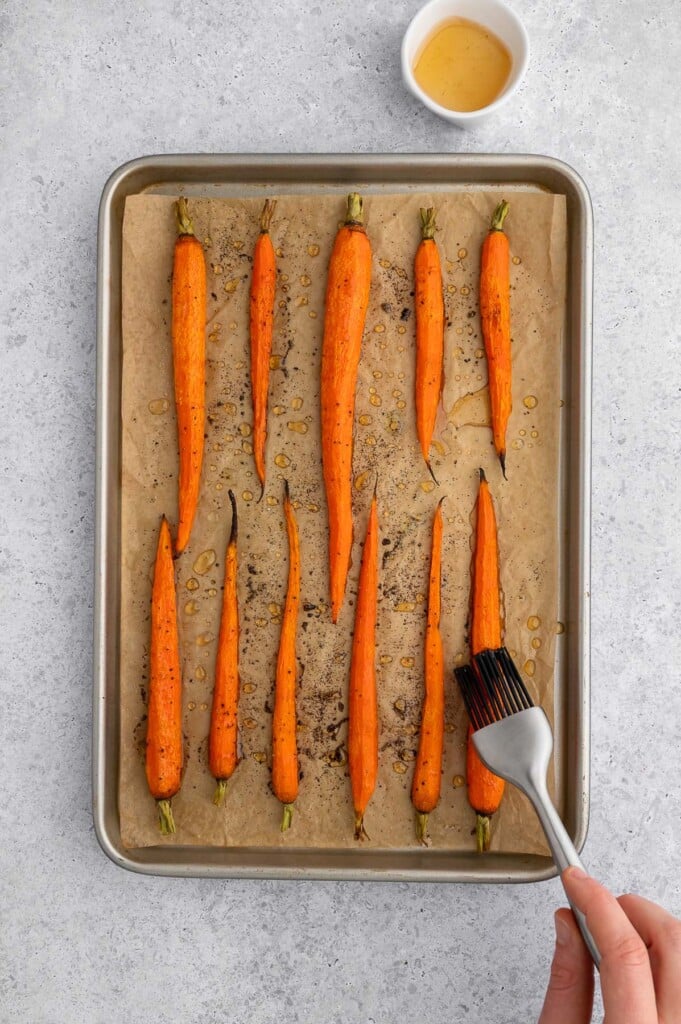 A pastry brush brushing a honey garlic glaze onto carrots on a parchment lined baking sheet.