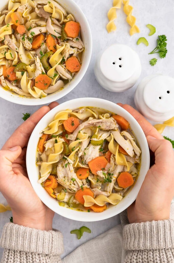 Two hands holding up a bowl of rotisserie chicken soup with another bowl of soup sitting on the marble counter in the background