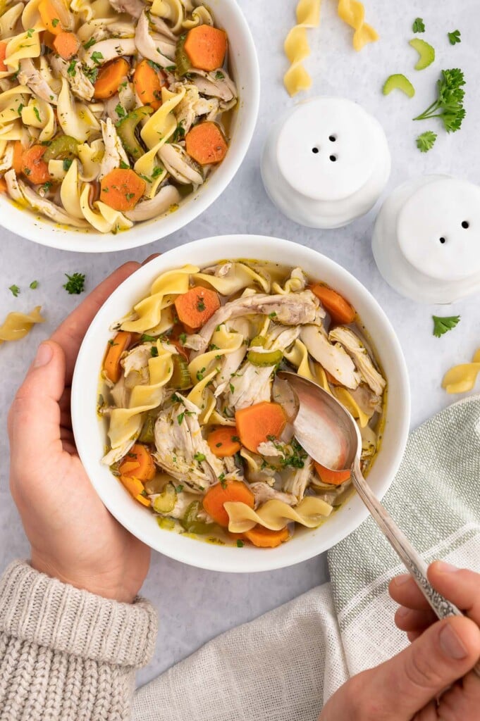 A hand holding up a white bowl of rotisserie chicken soup with a spoon dipping in it with another bowl of soup sitting on a marble counter