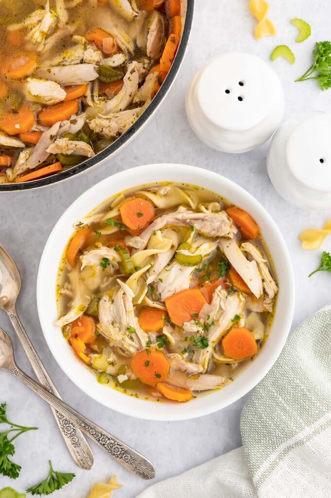 A white bowl of rotisserie chicken soup with the dutch oven of the soup on the side on a marble counter