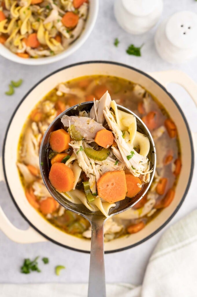 A ladle of rotisserie chicken soup holding up with a dutch oven in the background