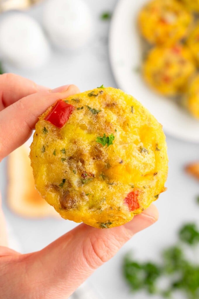 A hand holding up a sausage egg muffin with a plate of them in the background.