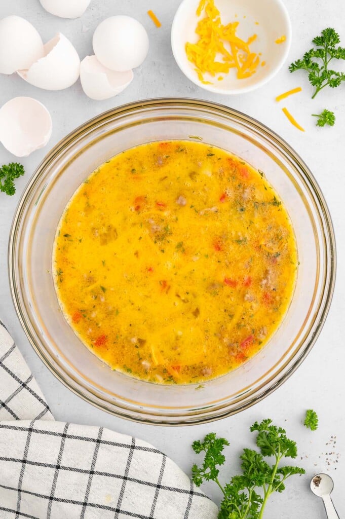 A clear bowl with whisked eggs, sausage, bell peppers, onions, and spices in it.