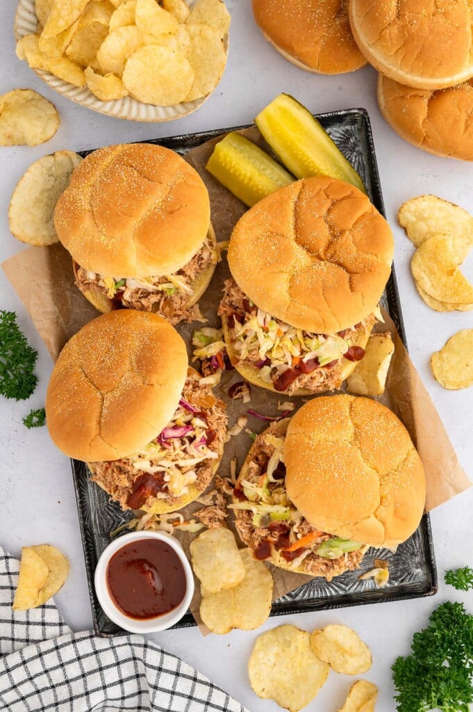 An overhead shot of four bbq chicken sandwiches on a platter with quartered pickles, a small bowl of bbq sauce and chips on the side.