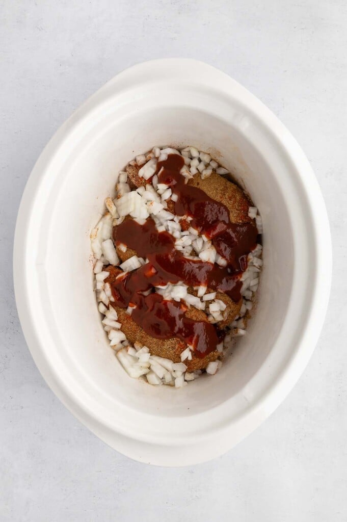 Uncooked chicken breasts, diced onions, dried seasonings, and bbq sauce in a white crockpot.