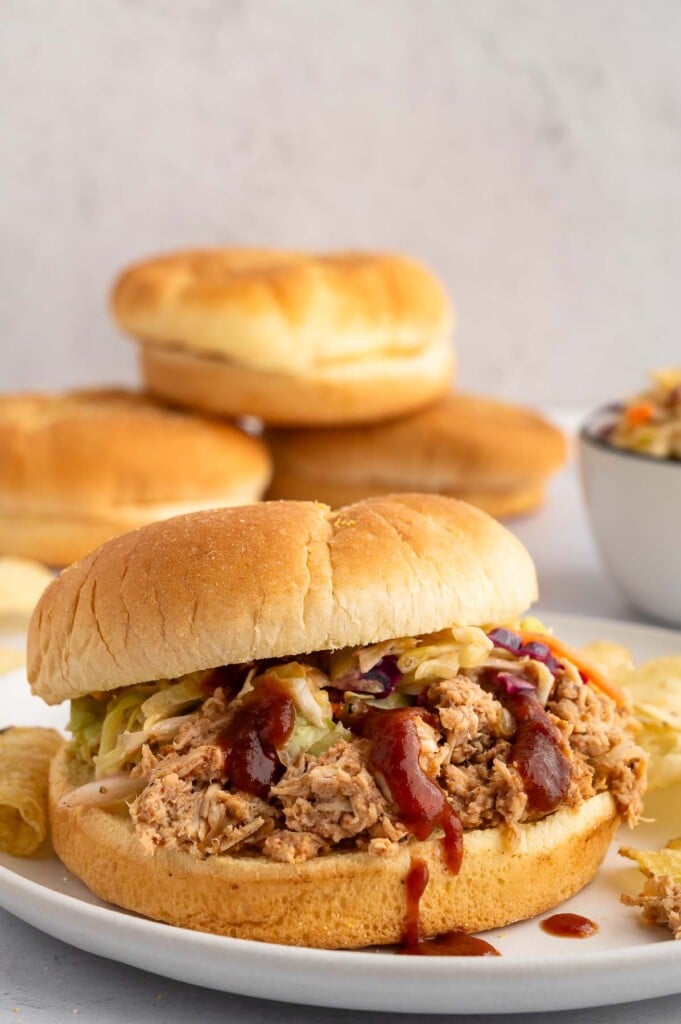 A Slow Cooker BBQ Chicken sandwich with cole slaw and bbq sauce on it. with more buns in the background.