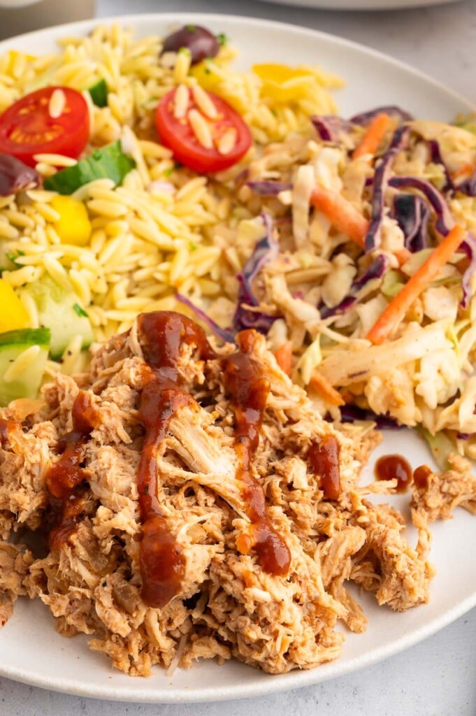 Slow Cooker BBQ Chicken on a white plate with cole slaw and orzo salad in the background.