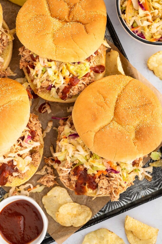 An overhead shot of 3 bbq chicken sandwiches on a platter with a small bowl of bbq sauce and chips on the side.