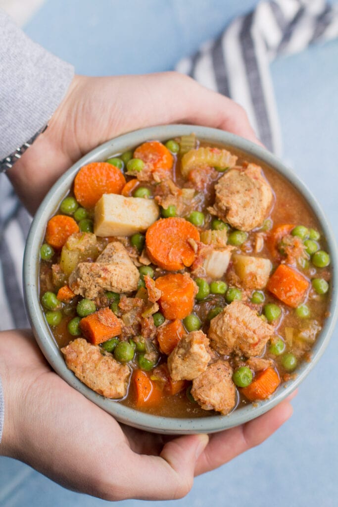 Two hands holding a bowl of crockpot chicken stew over a grey background.
