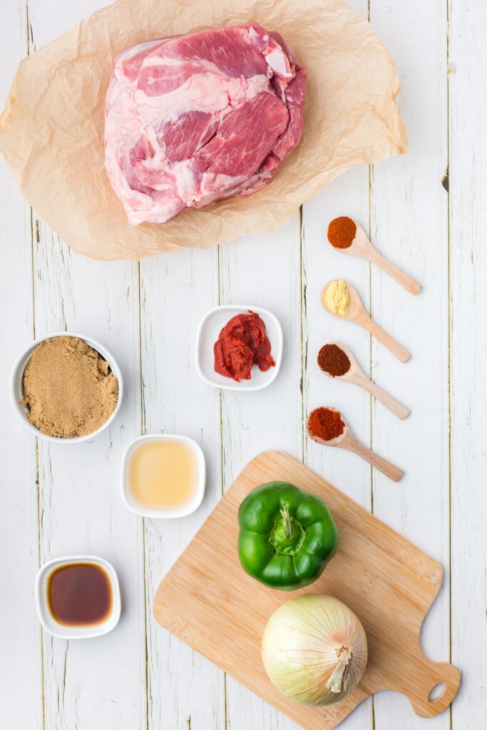 Ingredients for slow cooker pulled pork in white bowls: pork butt roast, onion, bell pepper, brown sugar, tomato paste, apple cider vinegar, chili powder, dry mustard, Worcestershire sauce, smoked paprika