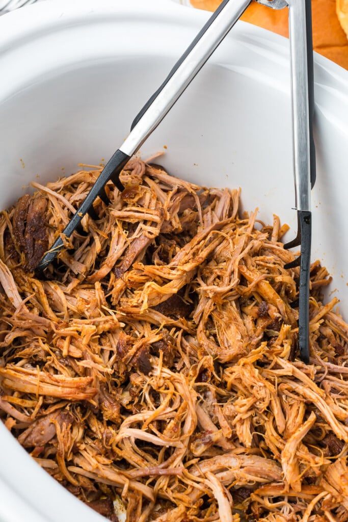 Slow Cooker pulled pork in a white crockpot after being shredded with open tongs in the crockpot