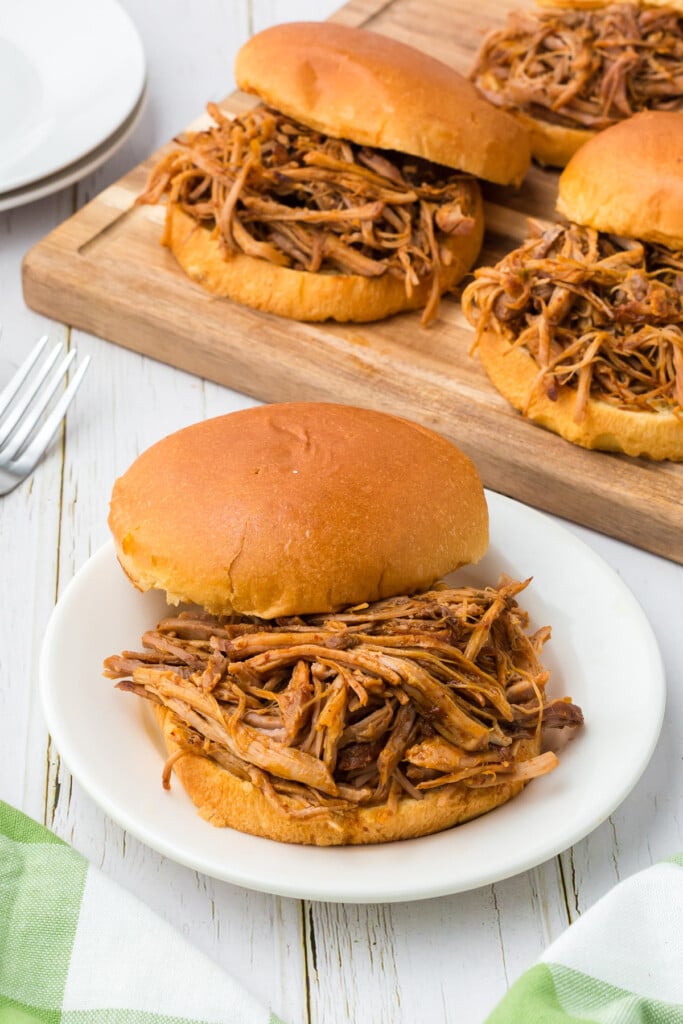 slow cooker pulled pork on a sandwich bun on a white plate with a wood platter of pulled pork sandwiches in the background