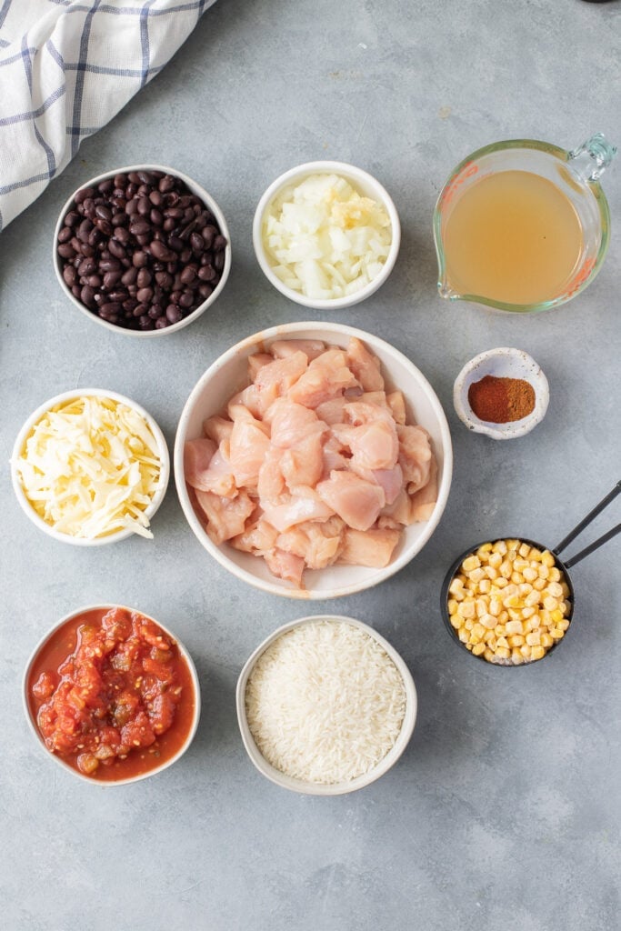 Ingredients for Southwest Chicken Casserole in small white bowls: white rice, minced garlic, chopped onion, chicken broth, chicken breasts, black beans, frozen corn, jarred salsa, chili powder, paprika, and cheddar cheese