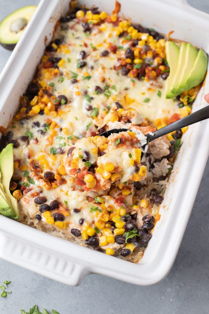 A black spoon scooping out a serving of healthy mexican chicken casserole with sliced avocados on the side