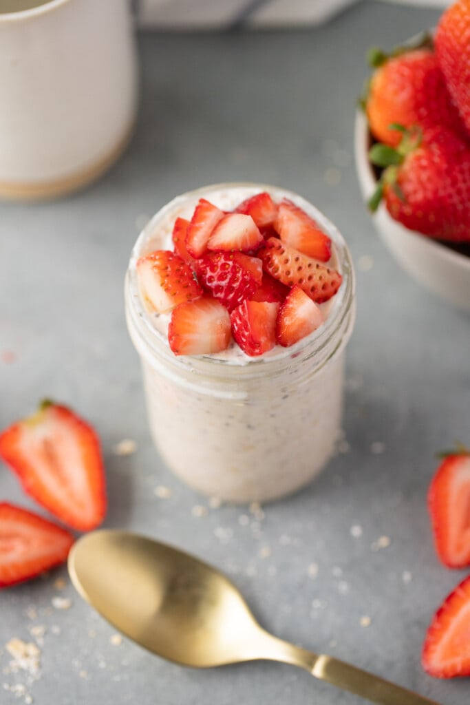 A mason jar of strawberry overnight oats on a grey background with a bowl of strawberries and a gold spoon on the side